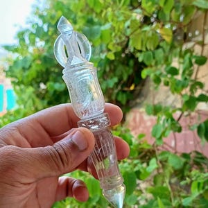 Op de afbeelding: Een helder, sierlijk glazen object met een puntige punt en een decoratieve bovenkant, tegen een achtergrond van groene bladeren. Het object heeft een gedetailleerd, gestructureerd ontwerp en lijkt een decoratief item of een spiritueel hulpmiddel te zijn.