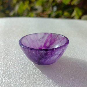 Può includere: Una piccola ciotola rotonda scolpita in pietra viola traslucida. L'interno e l'esterno della ciotola presentano un motivo a vortice di tonalità viola più chiare e più scure. La ciotola è appoggiata su una superficie bianca con uno sfondo verde sfocato.