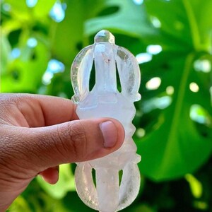 May include: A clear, carved crystal object with a detailed design, held in a hand against a backdrop of green foliage. The crystal has a symmetrical shape with rounded and pointed elements.