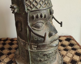 May include: A bronze sculpture of a human head with elaborate details and a textured surface. The head is adorned with two large, ornate ears and a geometric pattern on the forehead. The sculpture is a traditional African art piece.