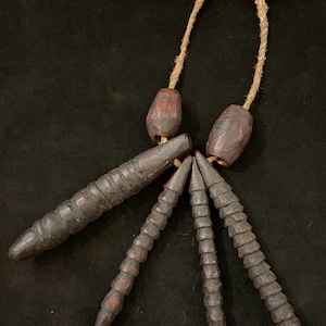 Könnte beinhalten: Eine handgefertigte, dunkelbraune Halskette mit einer natürlichen Schnur. Die Halskette hat mehrere geschnitzte Holzelemente, darunter zylindrische und konische Formen, mit roten Akzenten. Das Design deutet auf eine rustikale, tribale Ästhetik hin.