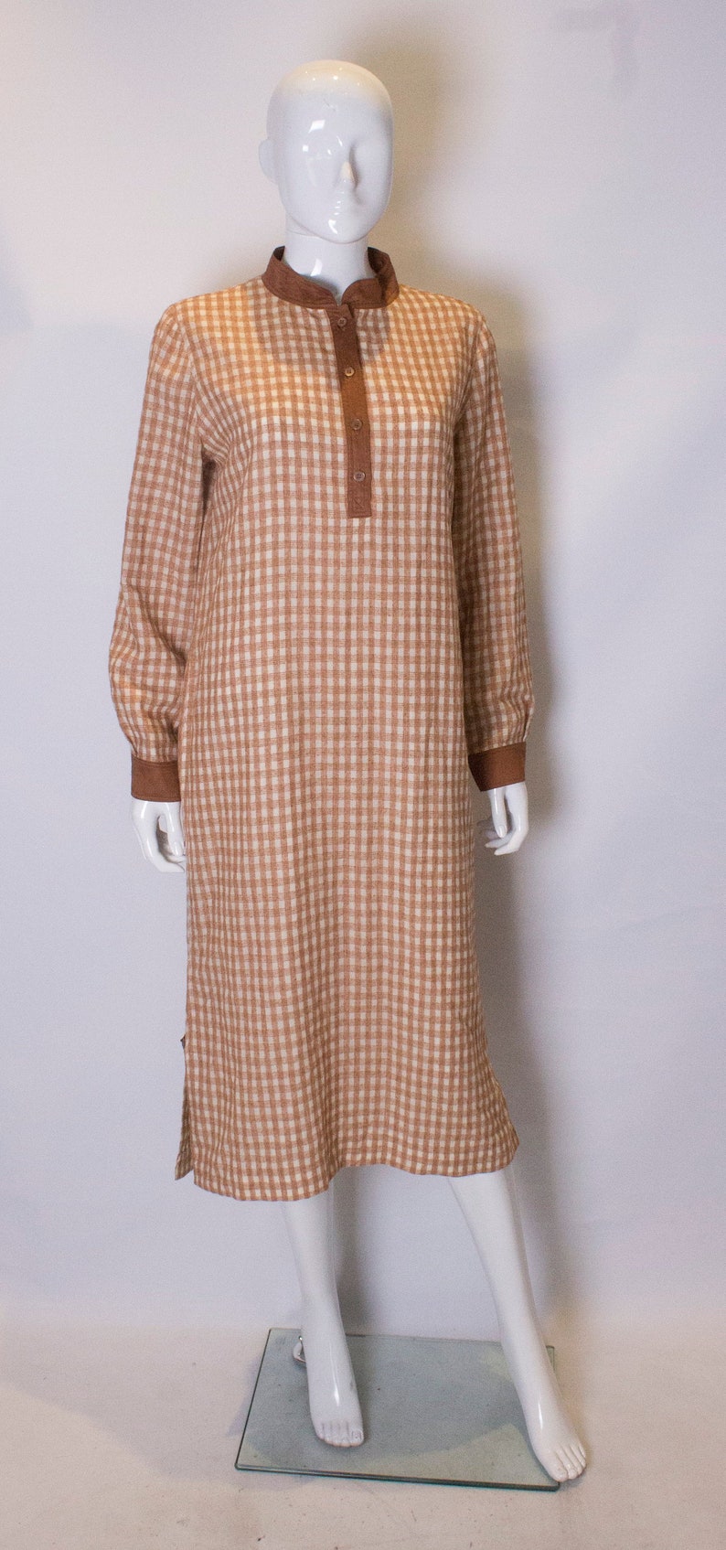 May include: A brown and white gingham dress with a brown collar and button closure. The dress has long sleeves and a knee-length hem.