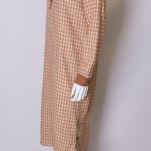 May include: A brown and white gingham dress with a brown collar and long sleeves. The dress has a button closure at the neck and a side slit.