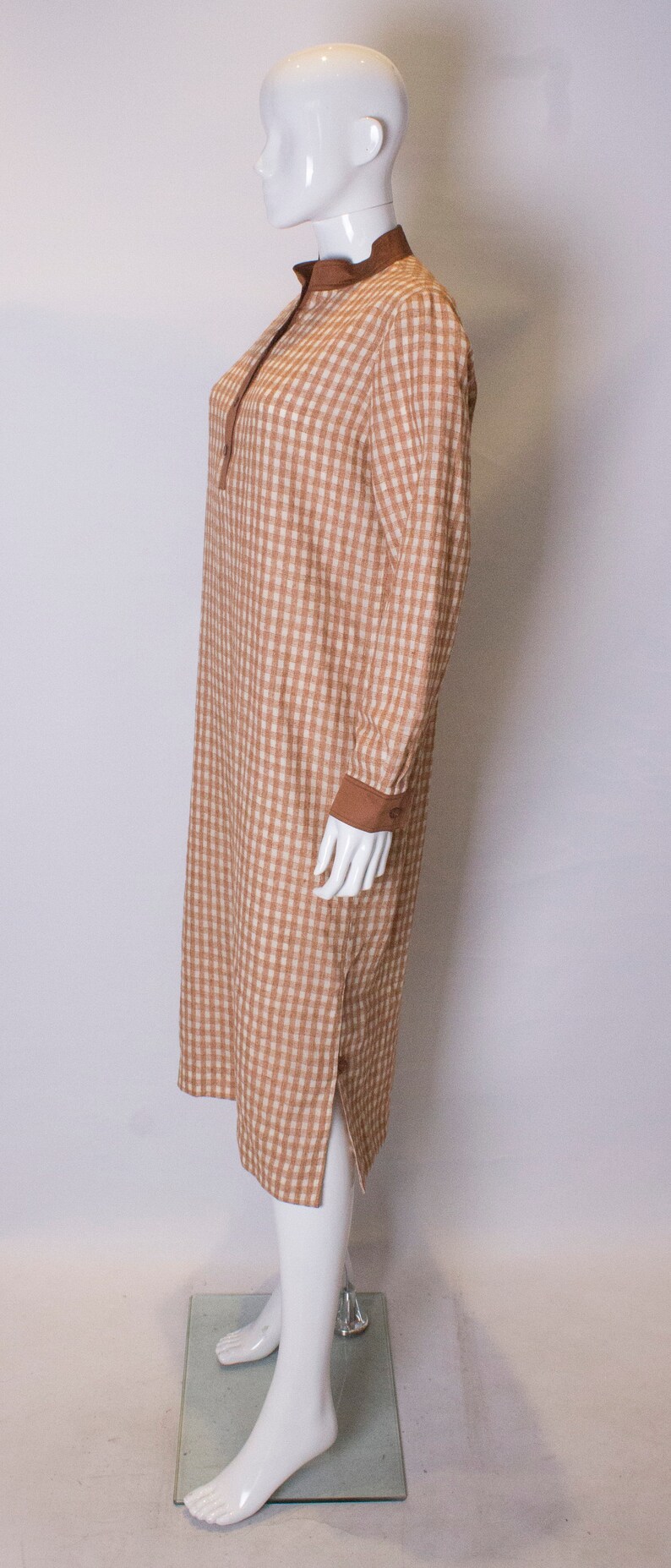 May include: A brown and white gingham dress with a brown collar and long sleeves. The dress has a button closure at the neck and a side slit.