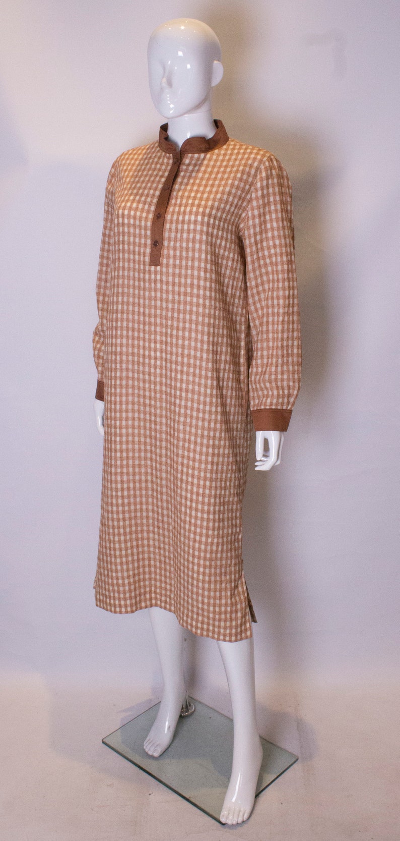 May include: A brown and white gingham dress with a brown collar and button closure. The dress has long sleeves and a loose fit.
