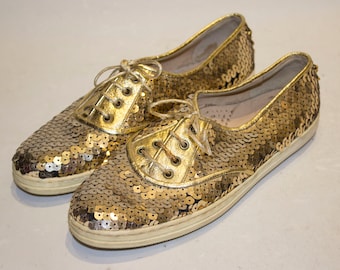 Könnte beinhalten: Ein Paar goldene Sneakers mit Pailletten und weißen Schnürsenkeln. Die Sneakers haben einen niedrigen Schaft und eine runde Spitze.