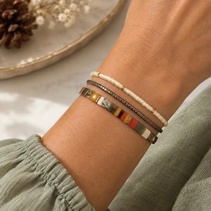 Puede incluir: Un conjunto de tres pulseras usadas juntas. La pulsera superior consiste en pequeñas cuentas de color crema con detalles dorados. La pulsera del medio presenta cuentas cilíndricas metálicas oscuras. La pulsera inferior tiene cuentas planas y cuadradas en un patrón de tonos rojos, amarillos, verdes, blancos y metálicos.