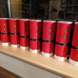 Może przedstawiać: Osiem czerwono-białych puszek świątecznych z czarnymi obw&oacute;dkami i złotymi akcentami. Puszki są ułożone w rzędzie na drewnianej powierzchni.