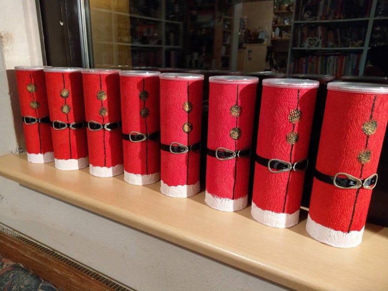 Może przedstawiać: Osiem czerwono-białych puszek świątecznych z czarnymi obw&oacute;dkami i złotymi akcentami. Puszki są ułożone w rzędzie na drewnianej powierzchni.