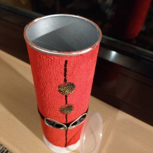 Może przedstawiać: Czerwono-biały kartonowy tubus z czarnym paskiem i złotymi kropkami. Tubus ma czarną i srebrną krawędź oraz przezroczysty plastikowy korek. Tubus jest ozdobiony tak, aby przypominał Świętego Mikołaja.