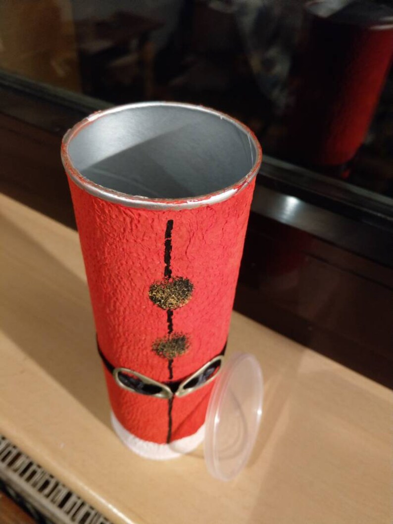 Może przedstawiać: Czerwono-biały kartonowy tubus z czarnym paskiem i złotymi kropkami. Tubus ma czarną i srebrną krawędź oraz przezroczysty plastikowy korek. Tubus jest ozdobiony tak, aby przypominał Świętego Mikołaja.