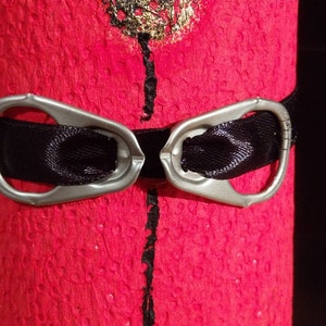 Może przedstawiać: Czerwona, fakturowana powierzchnia z czarną wstążką przewiązaną wok&oacute;ł. Do wstążki przymocowane są dwa srebrne uchwyty, tworząc element dekoracyjny.