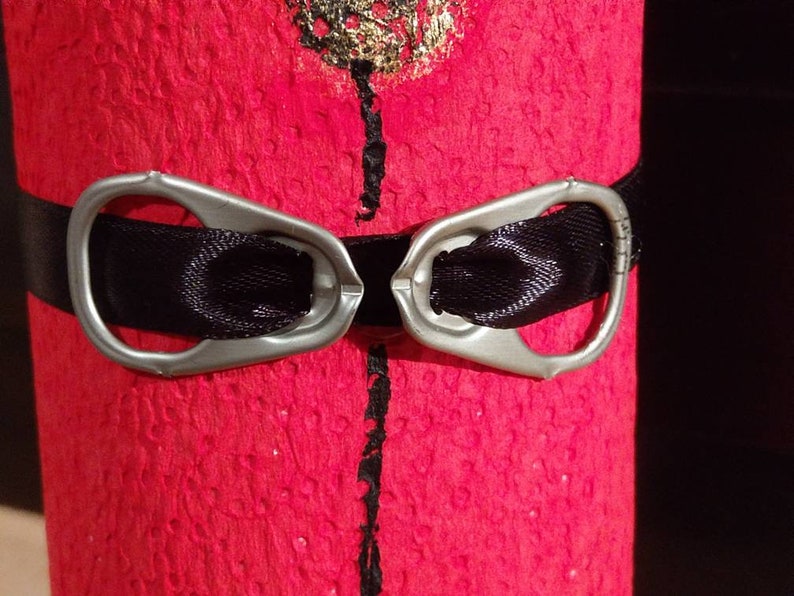 Może przedstawiać: Czerwona, fakturowana powierzchnia z czarną wstążką przewiązaną wok&oacute;ł. Do wstążki przymocowane są dwa srebrne uchwyty, tworząc element dekoracyjny.