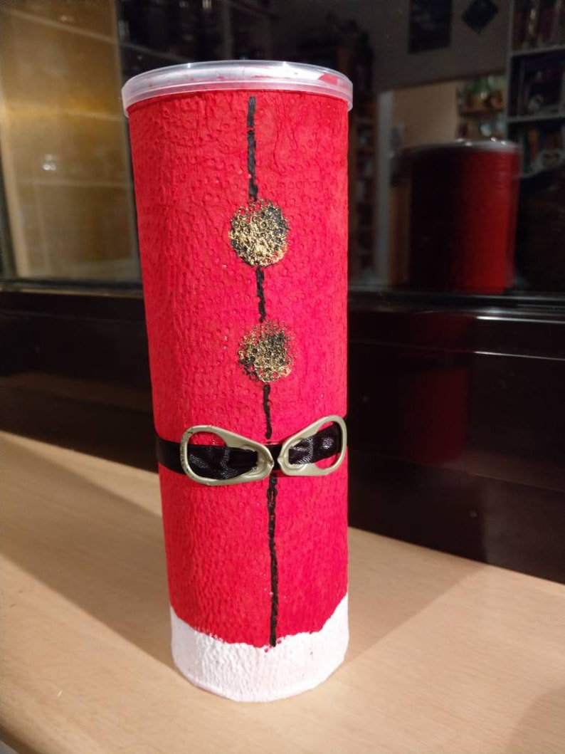 Może przedstawiać: Czerwony cylinder z białą podstawą, ozdobiony czarnymi liniami, złotymi k&oacute;łkami i czarną wstążką z dwoma srebrnymi uchwytami. Cylinder wykonany jest z fakturowanego materiału.