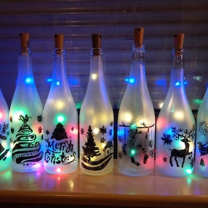 Könnte beinhalten: Acht gefrostete Glasflaschen mit Korkverschlüssen und bunten Lichtern im Inneren. Jede Flasche hat ein anderes Weihnachtsmotiv, darunter einen Weihnachtsbaum, ein Rentier und Schneeflocken.