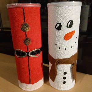 Może przedstawiać: Dwa kartonowe tuby ozdobione jako świąteczne postacie. Jeden jest pomalowany na czerwono z czarnym paskiem i białym wykończeniem, a drugi jest pomalowany na biało z brązowym szalikiem i czarnymi guzikami. Czerwony tubus ma złote k&oacute;łka z przodu.