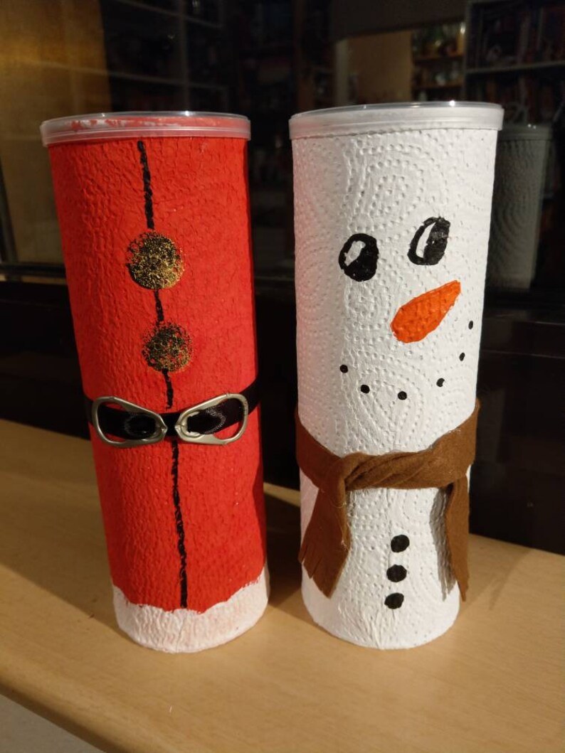 Może przedstawiać: Dwa kartonowe tuby ozdobione jako świąteczne postacie. Jeden jest pomalowany na czerwono z czarnym paskiem i białym wykończeniem, a drugi jest pomalowany na biało z brązowym szalikiem i czarnymi guzikami. Czerwony tubus ma złote k&oacute;łka z przodu.