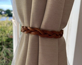 May include: A brown leather curtain tie with a braided design. The tie is holding back a pair of beige curtains with white stripes.