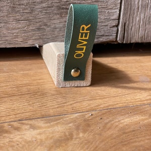 Puede incluir: Un tope de puerta de madera con un asa de cuero verde en forma de lazo. El asa tiene el nombre "OLIVER" grabado en letras doradas.