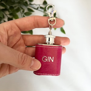 May include: A small, magenta leather flask with the word "GIN" in silver lettering. It has a silver cap and is attached to a silver keychain clip. The flask is designed to hold spirits.