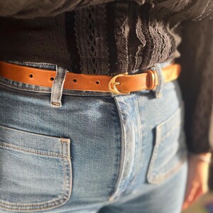 Könnte beinhalten: Ein hellbrauner Ledergürtel mit goldfarbener Schnalle wird mit einer blauen Jeans getragen. Der Gürtel wird um die Taille befestigt, und die Jeans hat Front- und Gesäßtaschen. Das Outfit wird durch ein schwarzes Oberteil ergänzt.