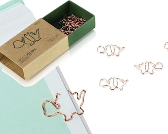 May include: A box of gold dinosaur-shaped paper clips. The box is made of brown cardboard with the words "Dino Clips" printed on it. The box is open and the clips are visible inside. There are also several clips scattered on a white surface.