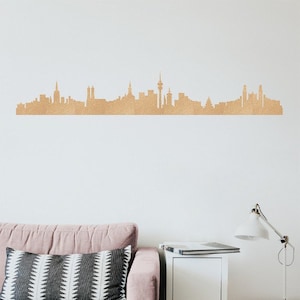 Könnte beinhalten: Goldfarbene Skyline-Silhouette einer Stadt, an einer weißen Wand befestigt. Die Skyline zeigt verschiedene Gebäudeformen und -höhen und bildet so ein detailliertes Stadtbild. Die Dekoration befindet sich über einem rosa Sofa mit einem gemusterten Kissen.