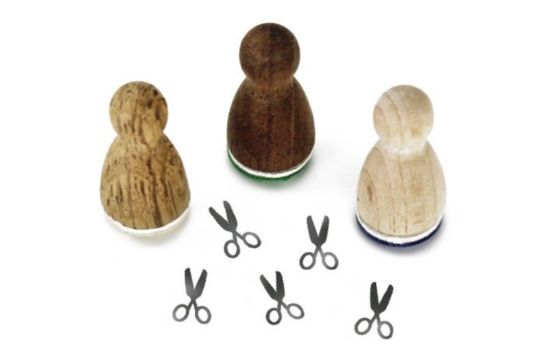 Könnte beinhalten: Drei Holzstempel in Form von Personen, mit unterschiedlichen Holzmaserungen. Jeder Stempel hat ein Tintenkissen in einer anderen Farbe. Fünf schwarze Scheren sind um die Stempel herum verstreut.