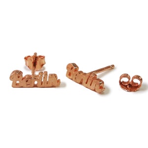 Könnte beinhalten: Ohrstecker aus Roségold mit dem Namen "Berlin" in Schreibschrift. Die Ohrringe werden auf einer weißen Oberfläche präsentiert, mit einem Ohrringstift und einem Schmetterlingsverschluss.