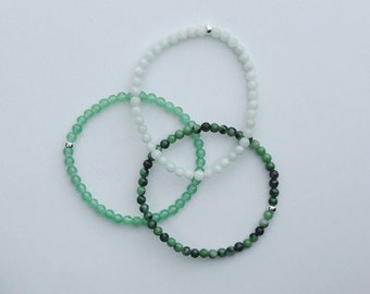 May include: Three beaded bracelets arranged in an overlapping circular pattern. One bracelet features pale mint green beads, one has translucent light green beads, and the third consists of dark forest green and mottled green beads, each with a silver bead.