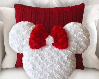 May include: A white plush pillow shaped like Minnie Mouse's head with a red bow. The pillow is sitting on a white couch with a red knitted pillow behind it.