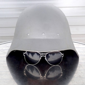 May include: A grey military helmet with a pair of black sunglasses resting on top. The helmet is sitting on a grey surface.