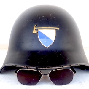 May include: A black helmet with a gold emblem featuring a hatchet and a shield with blue and white sections. Below the helmet are sunglasses with a silver frame and dark lenses. The helmet appears to be vintage.