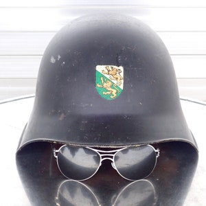 May include: A black helmet with a green and gold crest featuring two lions. The helmet has a visor and is sitting on a white surface. Sunglasses are resting on the helmet.