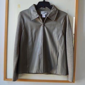 May include: A taupe-colored zip-up jacket featuring a smooth front panel and contrasting knit sleeves. The garment has a classic collar, a full-length center zipper, and a visible brand label on the inside neck area. The design is simple and functional.