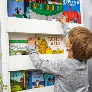 May include: A child reaches for a book titled "Rosie's Walk" on a white bookshelf. The bookshelf is filled with colorful children's books with titles like "Cat's Colors", "Adeline in London", and "Happy Monkey's Showbusiness".