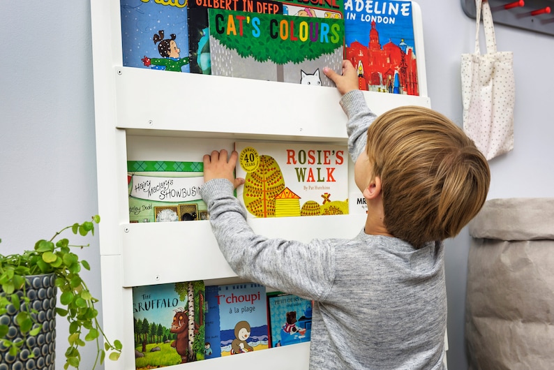 May include: A child reaches for a book titled "Rosie's Walk" on a white bookshelf. The bookshelf is filled with colorful children's books with titles like "Cat's Colors", "Adeline in London", and "Happy Monkey's Showbusiness".