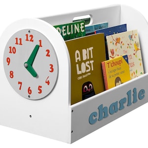 Könnte beinhalten: Weißer Holzbuchregals mit einem Zifferblatt auf der Vorderseite. Das Bücherregal hat einen grauen Boden mit dem Namen "charlie" in blauen Buchstaben. Das Bücherregal ist mit bunten Kinderbüchern gefüllt.