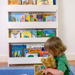 May include: A white bookshelf with four shelves filled with children's books. A young child is sitting on a blue mat holding a brown teddy bear and reading a book. The book titles include "Peace at Last", "Arthur's Tractor", "Little Miss Lucky", "The Bedtime Bear", "I Want My Mu", "Maisy's Bus", and "The Very Hungry Caterpillar".