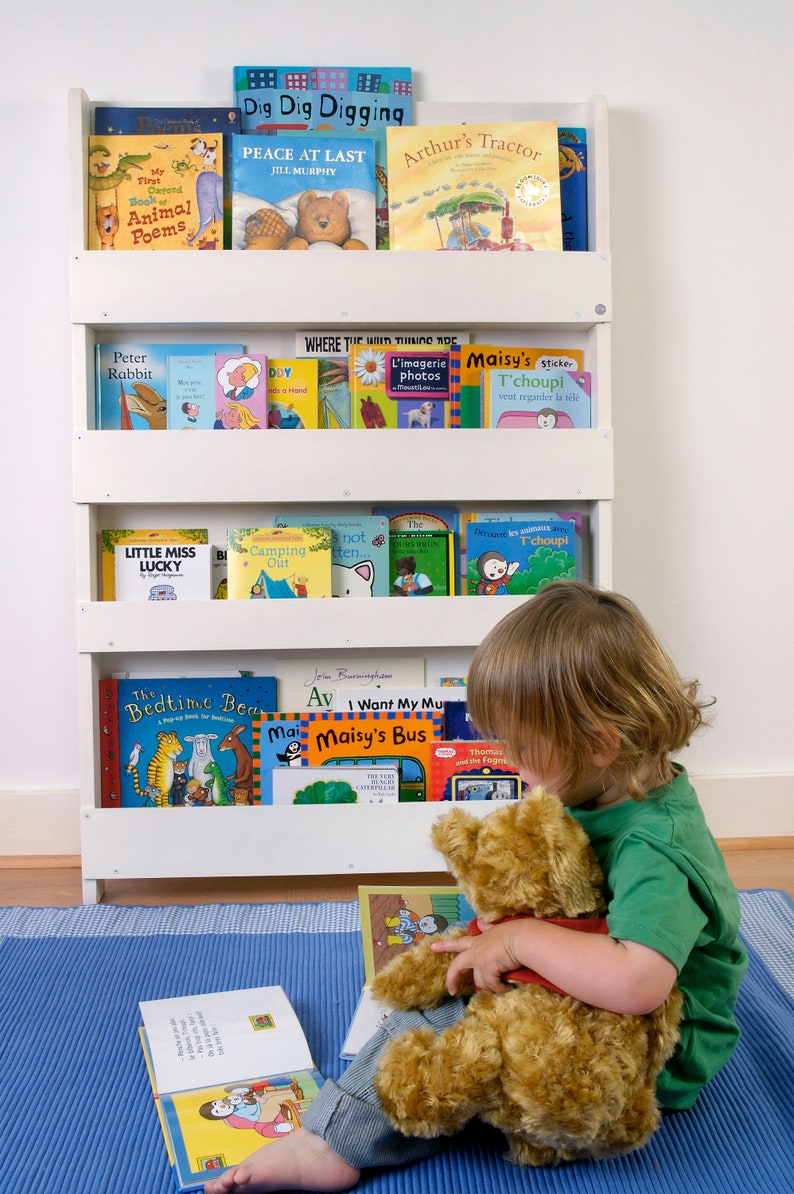 May include: A white bookshelf with four shelves filled with children's books. A young child is sitting on a blue mat holding a brown teddy bear and reading a book. The book titles include "Peace at Last", "Arthur's Tractor", "Little Miss Lucky", "The Bedtime Bear", "I Want My Mu", "Maisy's Bus", and "The Very Hungry Caterpillar".