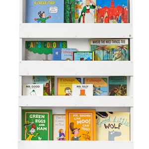 May include: White bookshelf with four shelves filled with children's books. The top shelf has books titled "Mimi Cracra", "Les Cent Metiers", "Inelecass", and "Madeline in London". The second shelf has books titled "Cat's Colors", "T'choupi a la plage", "Petit Ours Brun", and "Where the Wild Things Are". The third shelf has books titled "The Gruffalo", "Mr. Good", "T'choupi V'occupe bien de sa petite soeur", "Mr. Silly", "T'choupi joue de la musique", and "Rumpet Stiltskin". The bottom shelf has books titled "Green Eggs and Ham", "The Tiger Came to Tea", "Mr. Brown Can Moo! Can You?", and "Good Little Wolf".