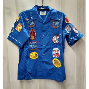May include: A bright blue short-sleeved button-down shirt featuring a pointed collar and numerous embroidered bowling patches. The patches display various text including AJBC, Brunswick, and 200, along with graphic bowling pins and balls.