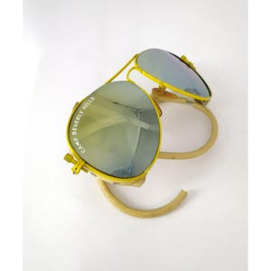 May include: Bright yellow sunglasses featuring reflective lenses with the text CAMP BEVERLY HILLS printed on the left lens. The frames have a distinct double bridge design and curved, light-colored temple arms that wrap around the lenses.