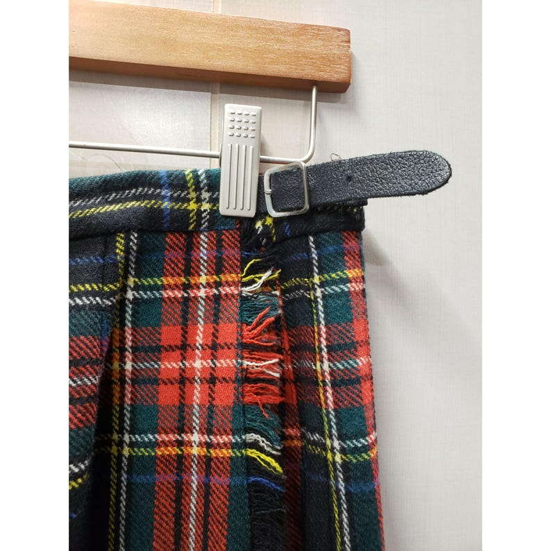 Op de afbeelding: Een close-up van een tartan kilt met een zwarte leren riem. De kilt heeft een klassiek ruitpatroon in rood, groen, geel en wit. Rafelige randen geven een vintage uitstraling. Opgehangen aan een houten kleerhanger.