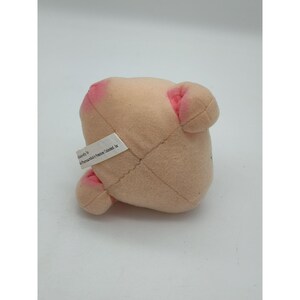 May include: A small, light beige plush toy with soft, rounded features and pink accents on the ears and snout. A white fabric tag is attached to the side, featuring black printed text that reads exclusively for Pharmaceuticals by Promotions Unlimited, Inc.