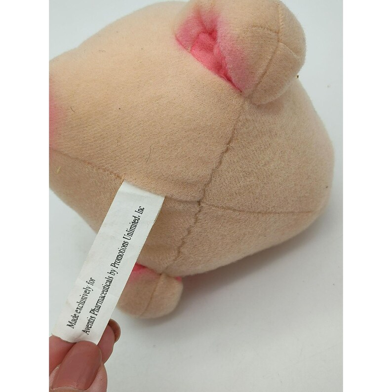 May include: A light beige plush toy with rounded ears featuring pink accents. A white fabric tag is attached to the side, printed with the text: Made exclusively for Aventis Pharmaceuticals by Promotions Unlimited, Inc.