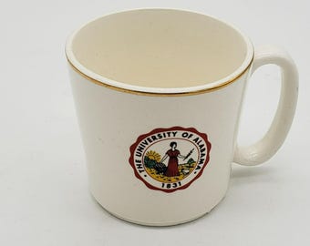 Könnte beinhalten: Cremefarbene Keramiktasse mit goldenem Rand und gebogenem Henkel. Die Tasse hat ein rundes Emblem mit den Worten "THE UNIVERSITY OF ALABAMA" und dem Jahr "1831". Das Emblem zeigt eine Frau in einem roten Kleid.