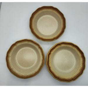 May include: Three round ceramic bowls with a speckled beige interior and a scalloped edge. Each bowl features a wide, dark brown band around the rim, creating a distinct contrast against the lighter, textured center of the stoneware dishware.