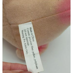May include: A tan and pink plush toy with a white fabric tag attached. The tag reads: Made in China, Content: polyester fiber, pellets, polyfoam, All new materials, WARNING: For Adults Only. The plush material has a soft, textured surface appearance.