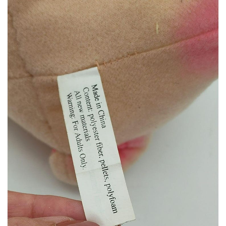 May include: A tan and pink plush toy with a white fabric tag attached. The tag reads: Made in China, Content: polyester fiber, pellets, polyfoam, All new materials, WARNING: For Adults Only. The plush material has a soft, textured surface appearance.